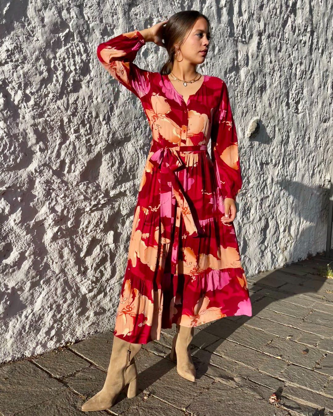 Mujer joven delante de pared blanca, vestida con vestido largo de otoño, en estampado en tonos rojos, de corte fluido, con vuelo y cinturón, de estilo bohemio, boho chic, de manga larga