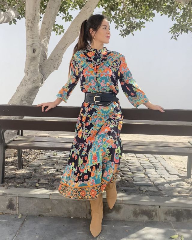 Mujer apoyada en banco de madera, vestida con conjunto de blusa y falda de otoño invierno en estampado floral multicolor sobre fondo negro de estilo folk