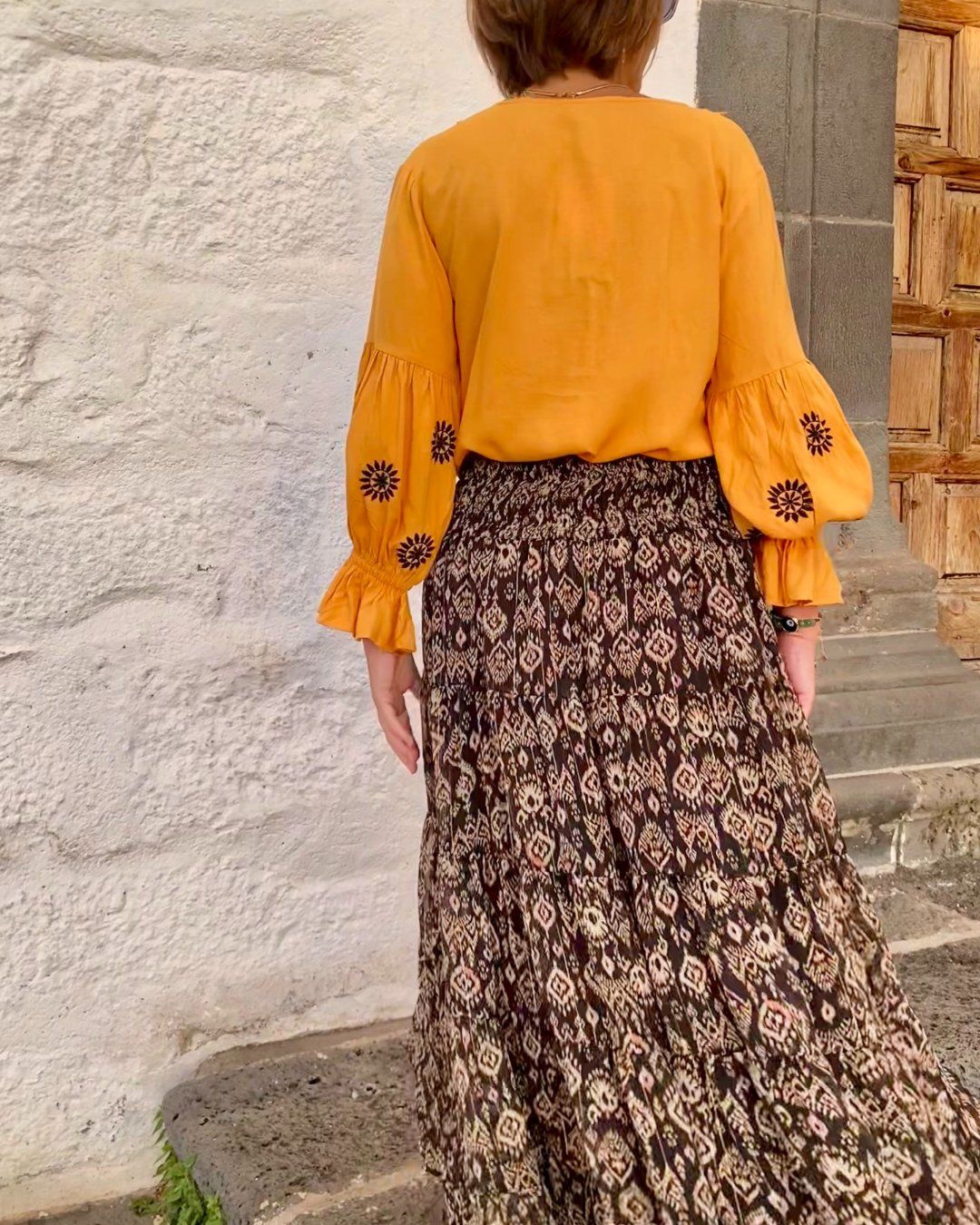 Imagen de espaldas de mujer vestida con blusa mostaza, de manga larga, con bordados en negro en las mangas globo. Lleva falda larga con estampados ikat, de gasa, en paneles, con cintura ancha con espejitos