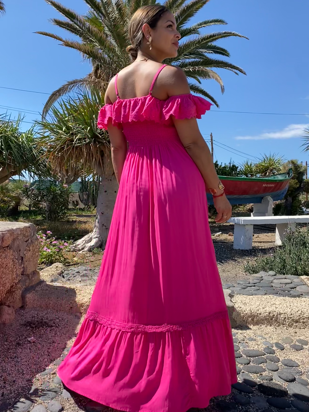 Mujer rubia con vestido largo fucsia de estilo ibicenco, con hombros descubiertos y tirantes. Escote ligeramente corazón con volantito de shifly que rodea los brazos. Busto elástico nido de abeja. De espaldas