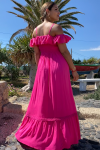 Mujer rubia con vestido largo fucsia de estilo ibicenco, con hombros descubiertos y tirantes. Escote ligeramente corazón con volantito de shifly que rodea los brazos. Busto elástico nido de abeja. De espaldas