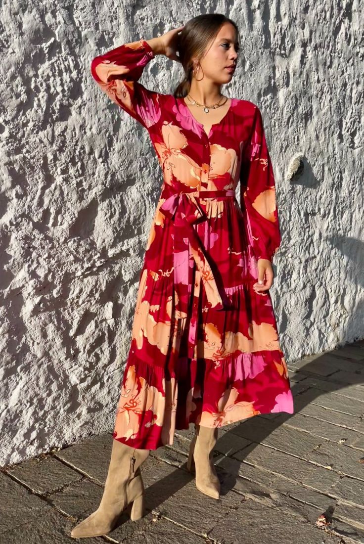 Mujer joven delante de pared blanca, vestida con vestido largo de otoño, en estampado en tonos rojos, de corte fluido, con vuelo y cinturón, de estilo bohemio, boho chic, de manga larga