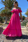 Mujer rubia con vestido largo fucsia de estilo ibicenco, con hombros descubiertos y tirantes. Escote ligeramente corazón con volantito de shifly que rodea los brazos. Busto elástico nido de abeja.