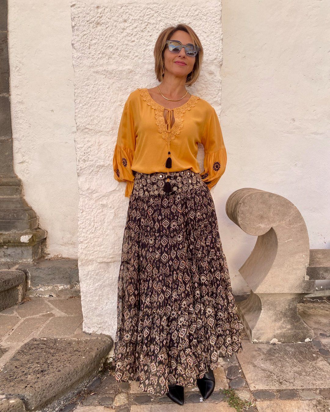 Mujer vestida con blusa mostaza, de manga larga, con bordados en negro en las mangas globo. Lleva falda larga con estampados ikat, de gasa, en paneles, con cintura ancha con espejitos