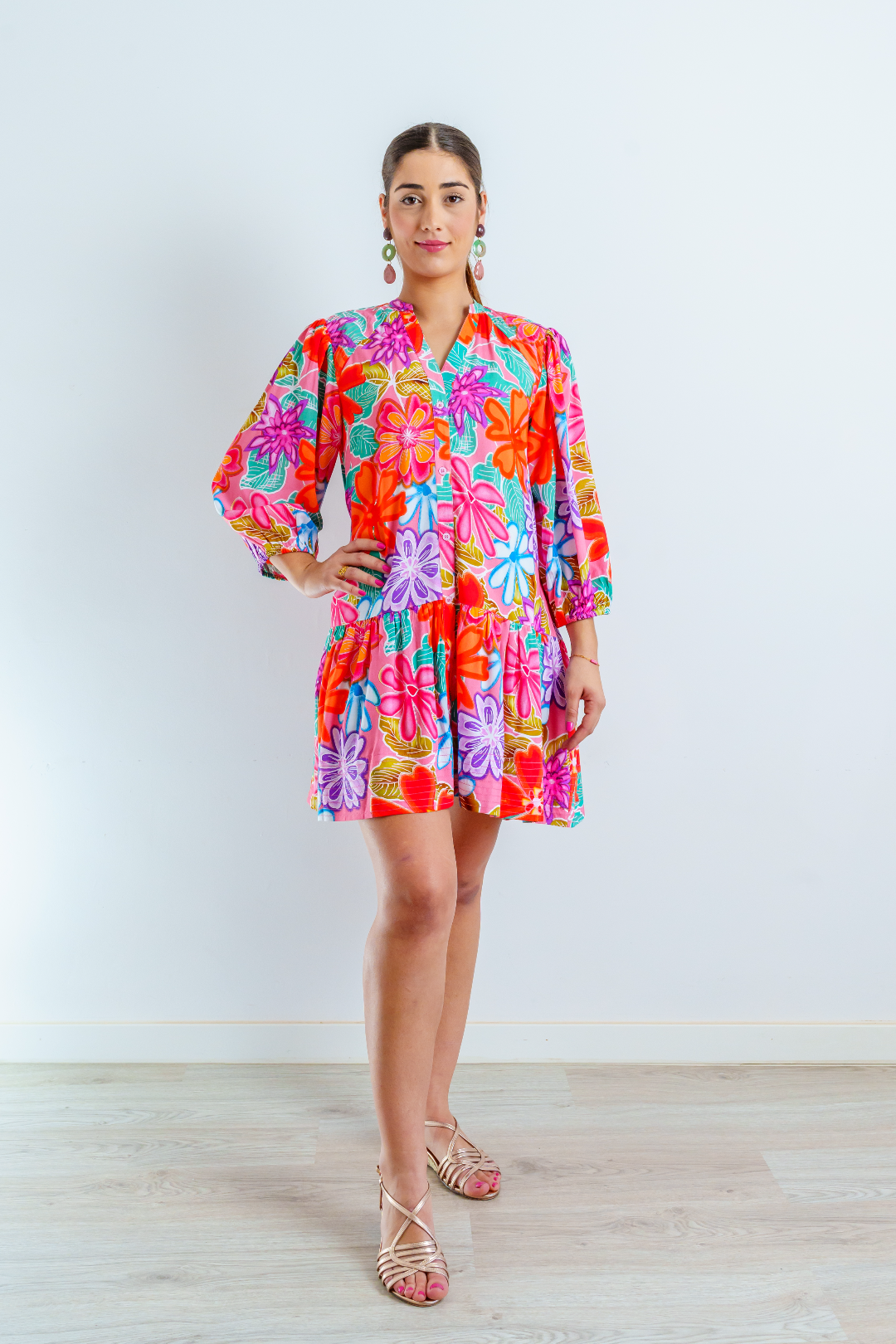 Imagen de cuerpo entero de mujer joven morena, con vestido corto en estampado floral multicolor sobre color rosa chicle. El vestido es abotonado con cuello Mao. Las mangas abullonadas de largo ¾ con elástico. El vestido es suelto y fluido.