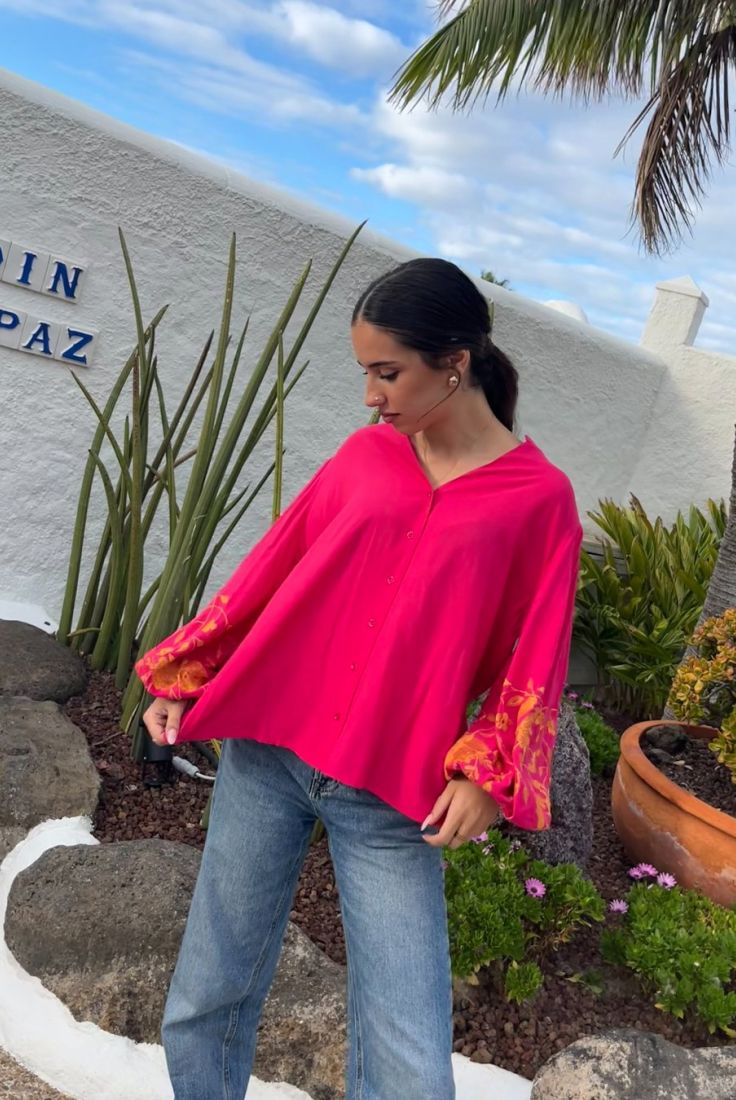 Mujer morena vestida con blusa fluida abotonada de color fucsia, con manga larga. Mangas abullonadas con bordados florales en color mostaza.