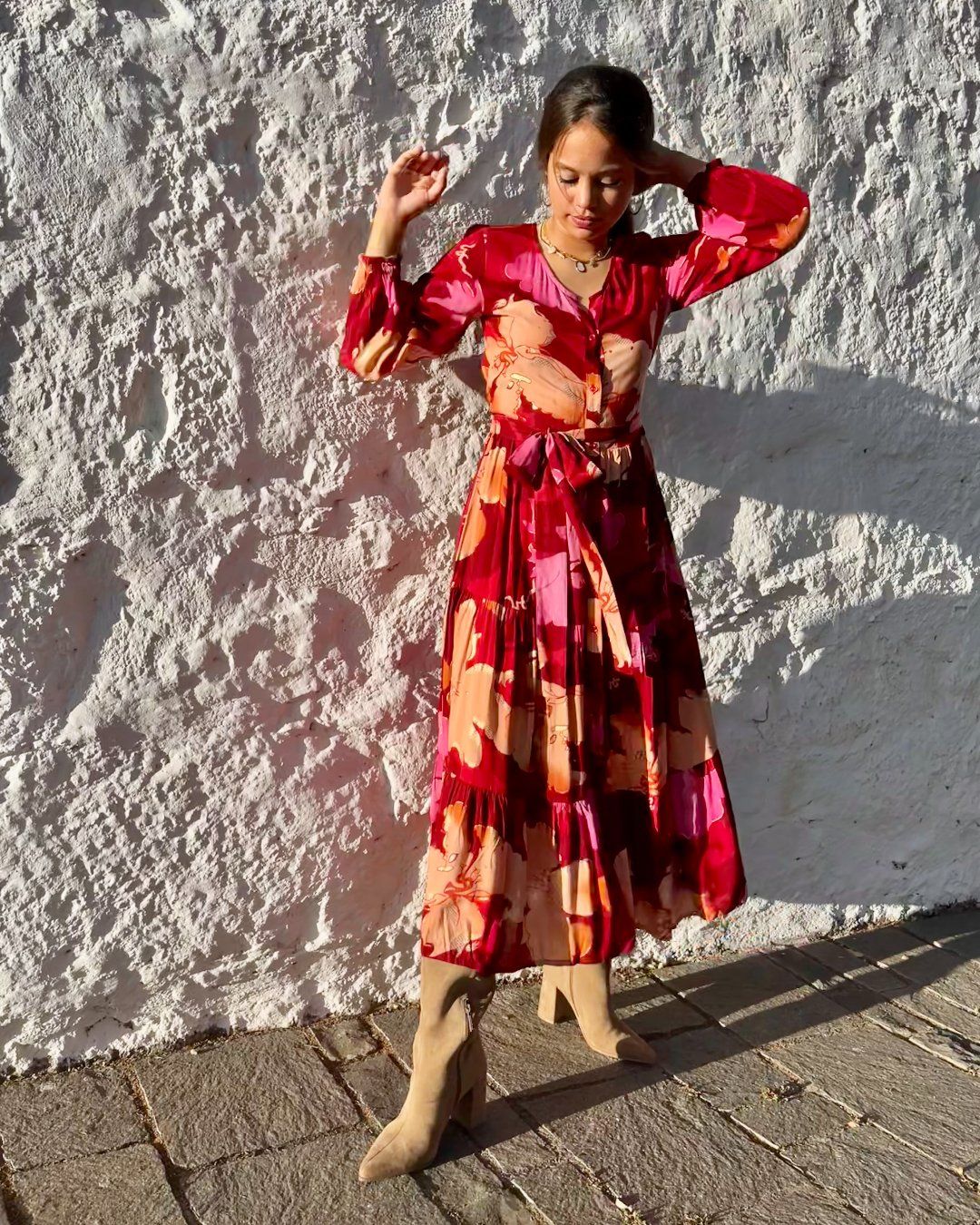Mujer joven delante de pared blanca, vestida con vestido largo de otoño, en estampado en tonos rojos, de corte fluido, con vuelo y cinturón, de estilo bohemio, boho chic, de manga larga