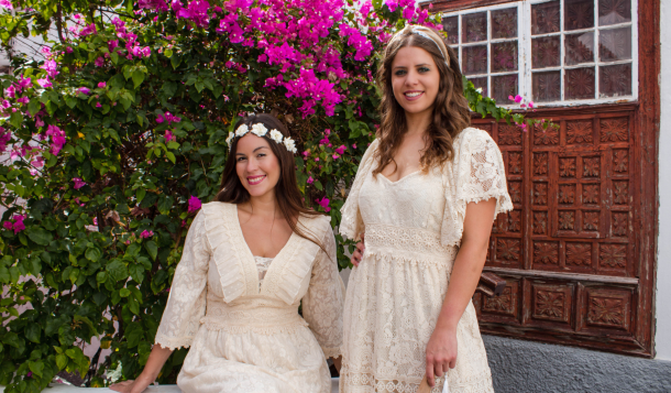 Imagen de dos mujeres vestidas con ropa para Los Indianos de La Palma, de color beige con encaje, de estilo bohemio delante de una planta de bouganvilla en Garachico