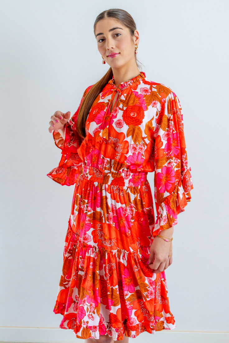 Imagen de medio cuerpo superior de mujer joven con vestido corto en estampado floral multicolor, en tonos rojos, sobro fondo blanco, con mangas 3/4 acampanadas, acabadas en volante.