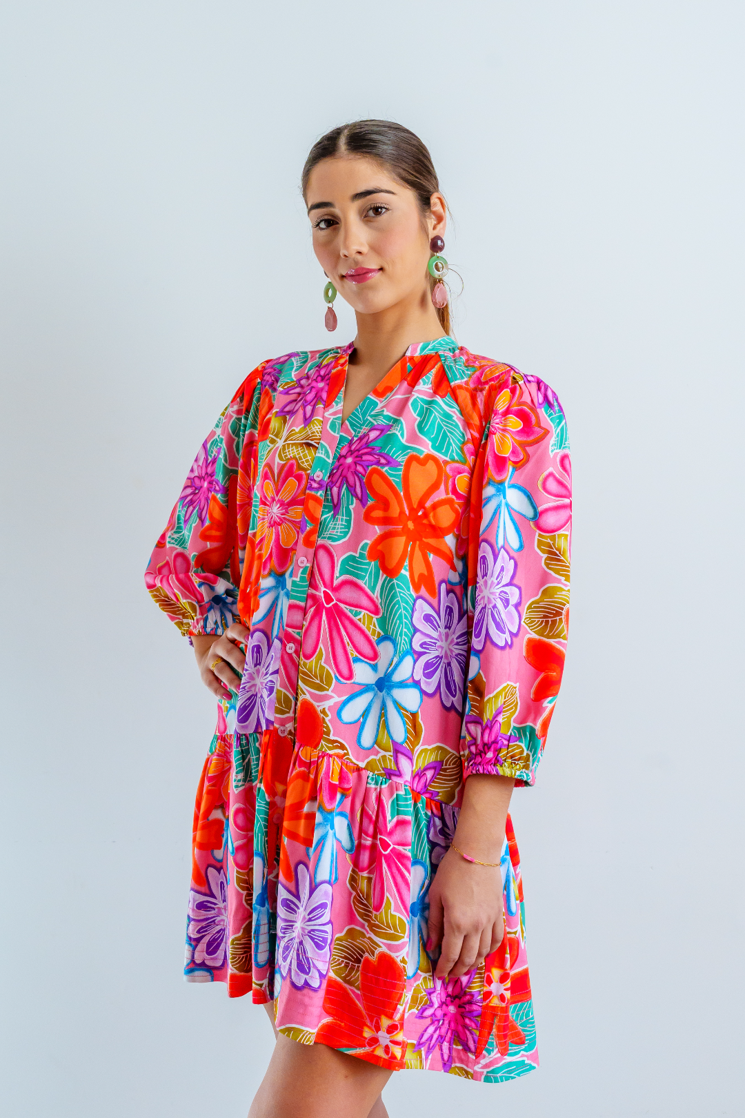 Imagen de medio cuerpo delantero de mujer joven morena, con vestido corto en estampado floral multicolor sobre color rosa chicle. El vestido es abotonado con cuello Mao. Las mangas abullonadas de largo ¾ con elástico. El vestido es suelto y fluido.