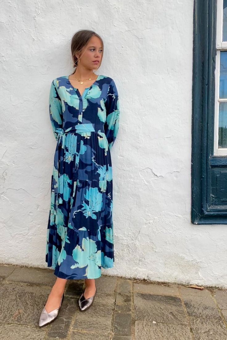 Mujer joven apoyada en pared blanca, vestida con vestido largo de otoño, en estampado en tonos azules, de corte fluido, con vuelo y cinturón, de estilo bohemio, boho chic, de manga larga
