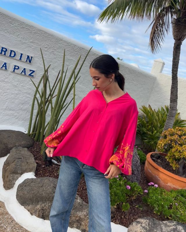 Mujer morena vestida con blusa fluida abotonada de color fucsia, con manga larga. Mangas abullonadas con bordados florales en color mostaza.