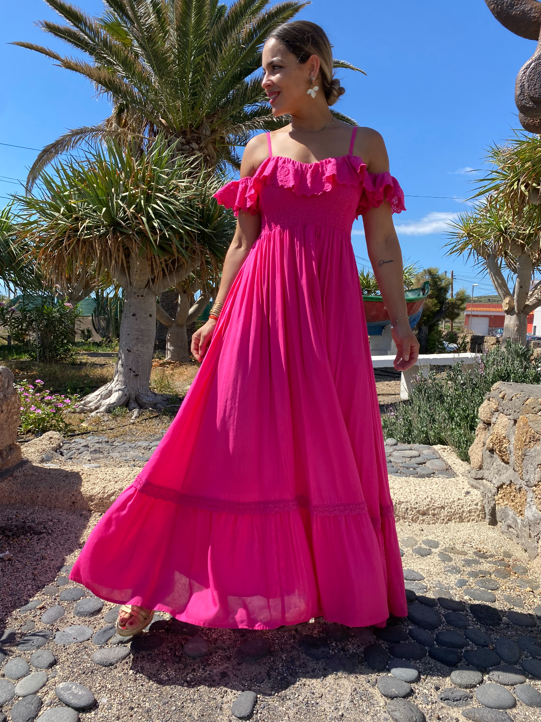 Mujer rubia con vestido largo fucsia de estilo ibicenco, con hombros descubiertos y tirantes. Escote ligeramente corazón con volantito de shifly que rodea los brazos. Busto elástico nido de abeja.