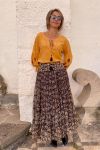 Mujer vestida con blusa mostaza, de manga larga, con bordados en negro en las mangas globo. Lleva falda larga con estampados ikat, de gasa, en paneles, con cintura ancha con espejitos