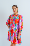 Imagen de medio cuerpo delantero de mujer joven morena, con vestido corto en estampado floral multicolor sobre color rosa chicle. El vestido es abotonado con cuello Mao. Las mangas abullonadas de largo ¾ con elástico. El vestido es suelto y fluido.