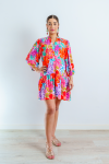 Imagen de cuerpo entero de mujer joven morena, con vestido corto en estampado floral multicolor sobre color rosa chicle. El vestido es abotonado con cuello Mao. Las mangas abullonadas de largo ¾ con elástico. El vestido es suelto y fluido.